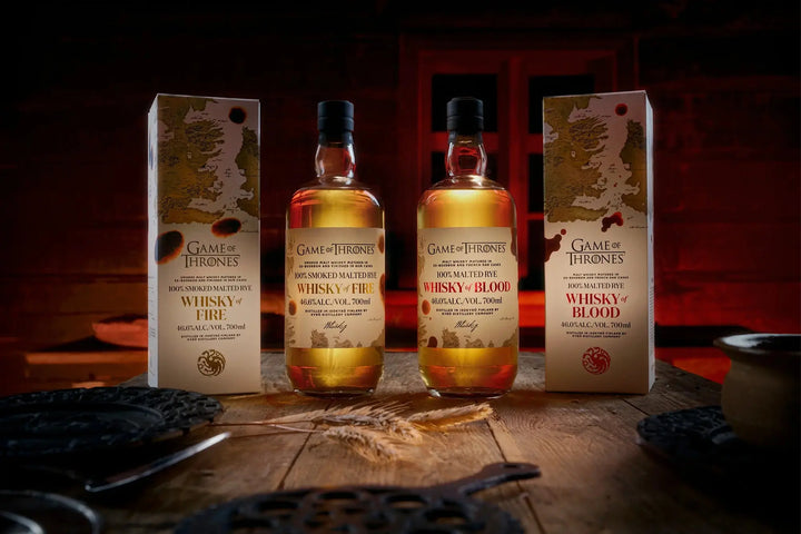 Three bottles of the Kyrö x Game of Thrones Whisky of Fire &amp; Blood Bundle (PRE-ORDER) from Main Street Liquor stand between matching boxes on a rustic wooden table, set against a dramatic, red-lit backdrop.