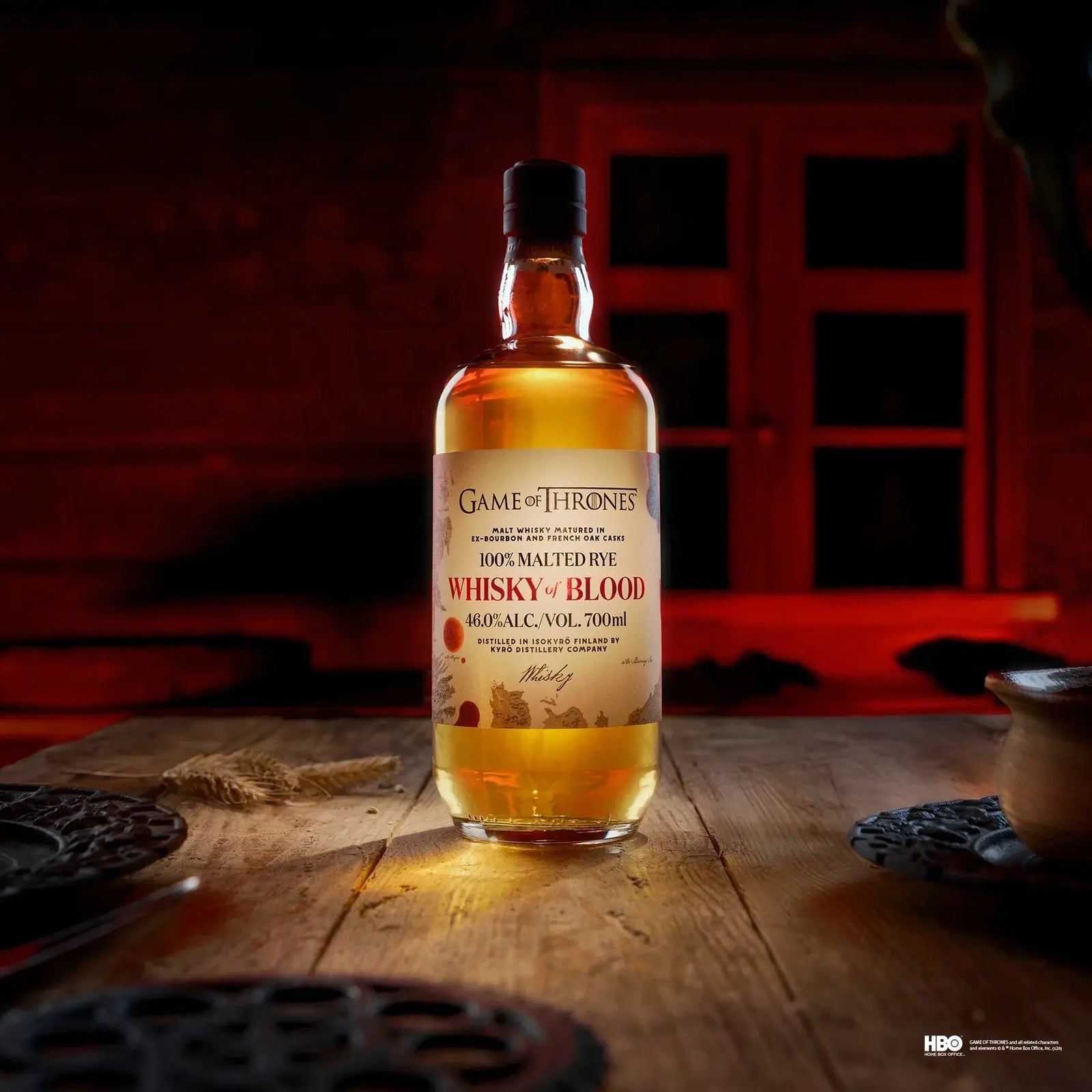 A bottle of Kyrö x Game of Thrones Whisky of Blood 700mL (PRE-ORDER) by Kyro Distillery sits on a wooden table in a dimly lit, red-glowing room, surrounded by plates and cups, with a window visible in the background.