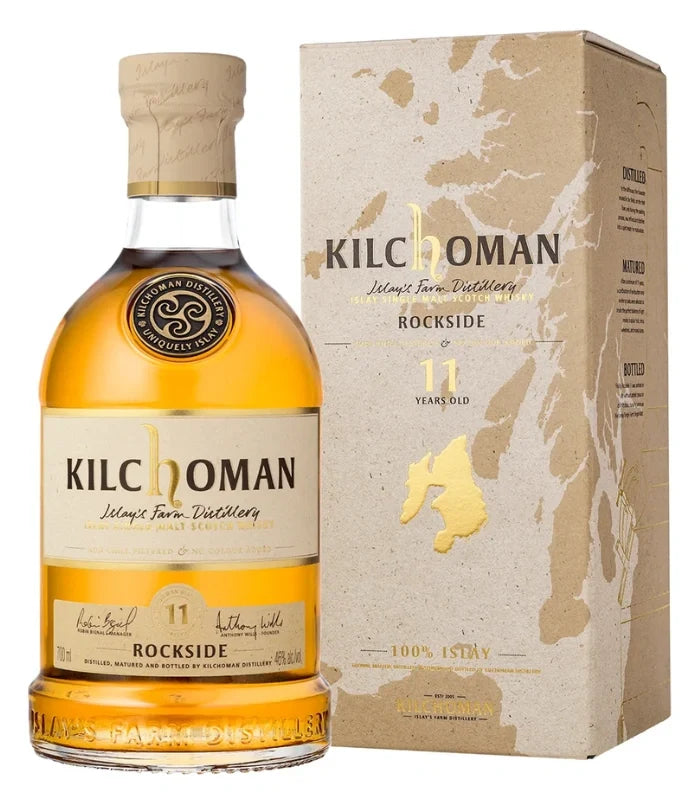 A bottle of Kilchoman Rockside 11 Year Single Malt Scotch Whisky 700mL with a tan and gold label, shown beside its matching box featuring a gold map outline and Kilchoman branding.
