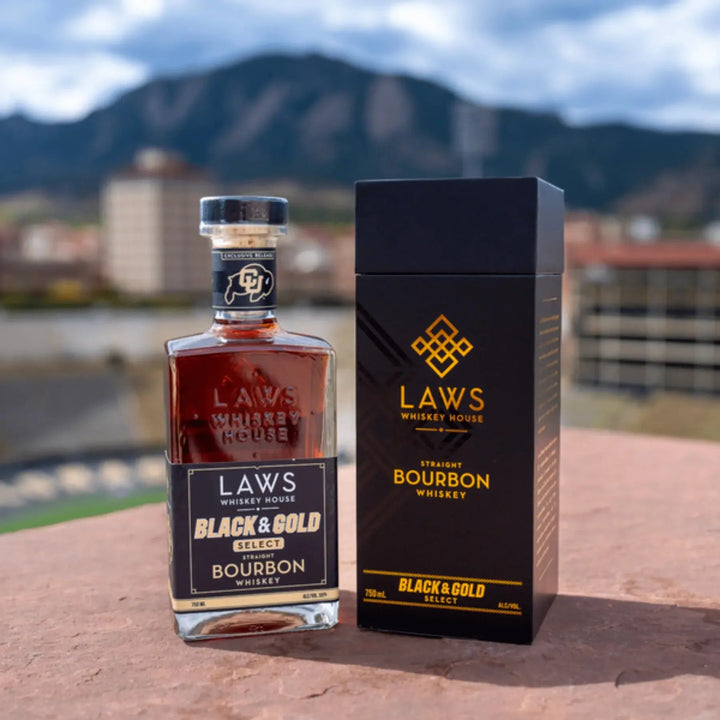 A bottle of Laws Whiskey House Black &amp; Gold Select Four Grain Straight Bourbon Whiskey stands beside its elegant black and gold box from Laws Whiskey House, with a blurred cityscape and mountain backdrop.