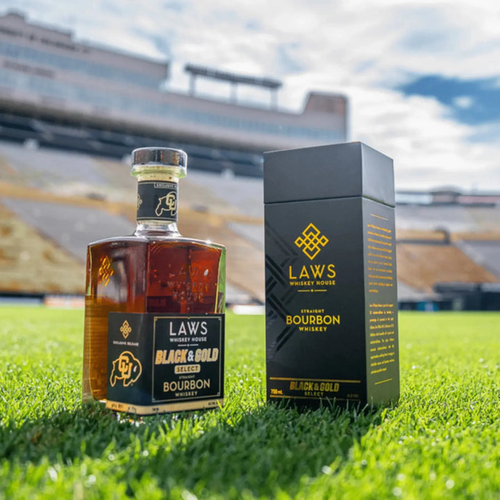 A bottle and box of Laws Whiskey House Black &amp; Gold Select Four Grain Straight Bourbon Whiskey rest on lush green grass, with a stadium and bleachers softly blurred in the background beneath a blue sky—showcasing this limited-release whiskey from Laws Whiskey House, crafted with their signature Colorado Four Grain Bourbon.
