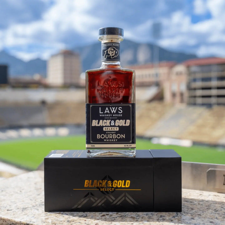 A bottle of Laws Whiskey House Black &amp; Gold Select Four Grain Straight Bourbon Whiskey from Laws Whiskey House, a limited-release expression, rests on its box against the backdrop of a blurred stadium and the majestic Colorado mountains beneath a partly cloudy sky.