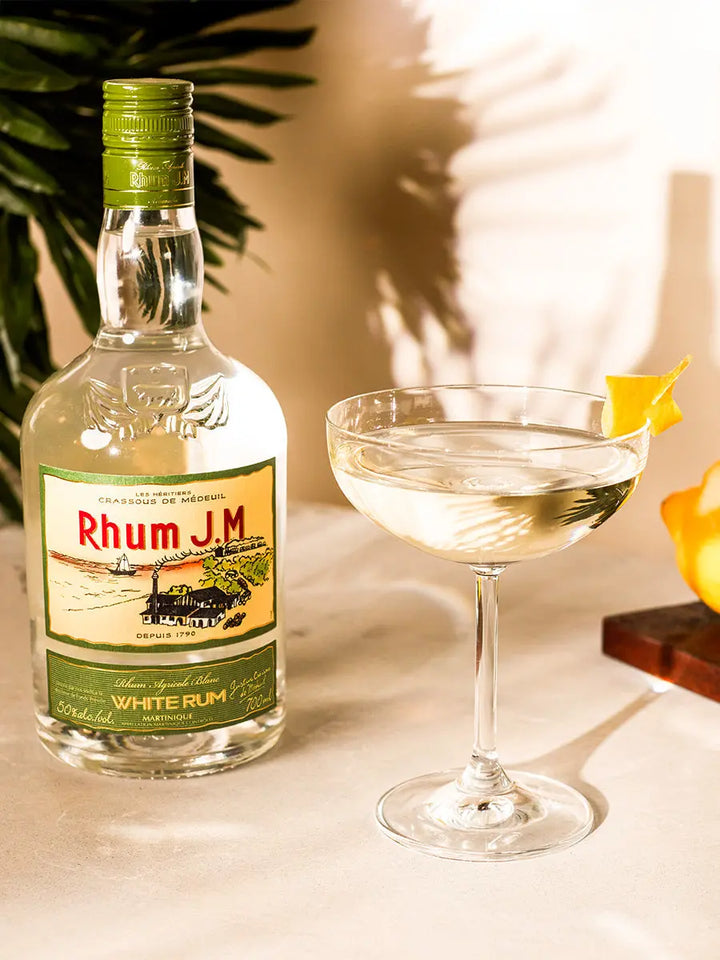 A bottle of Rhum J.M. Agricole Blanc 50 White Rum sits beside a coupe glass with a clear cocktail and lemon twist, while a leafy plant and lemon complete the vibrant scene.