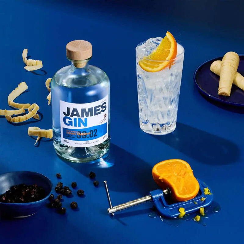 A bottle of James Gin Navy Strength 700mL (114 Proof) with a souvenir key chain stands next to a gin and tonic garnished with orange on a bright blue surface, surrounded by parsnip peels, juniper berries, and an orange in a blue vise.