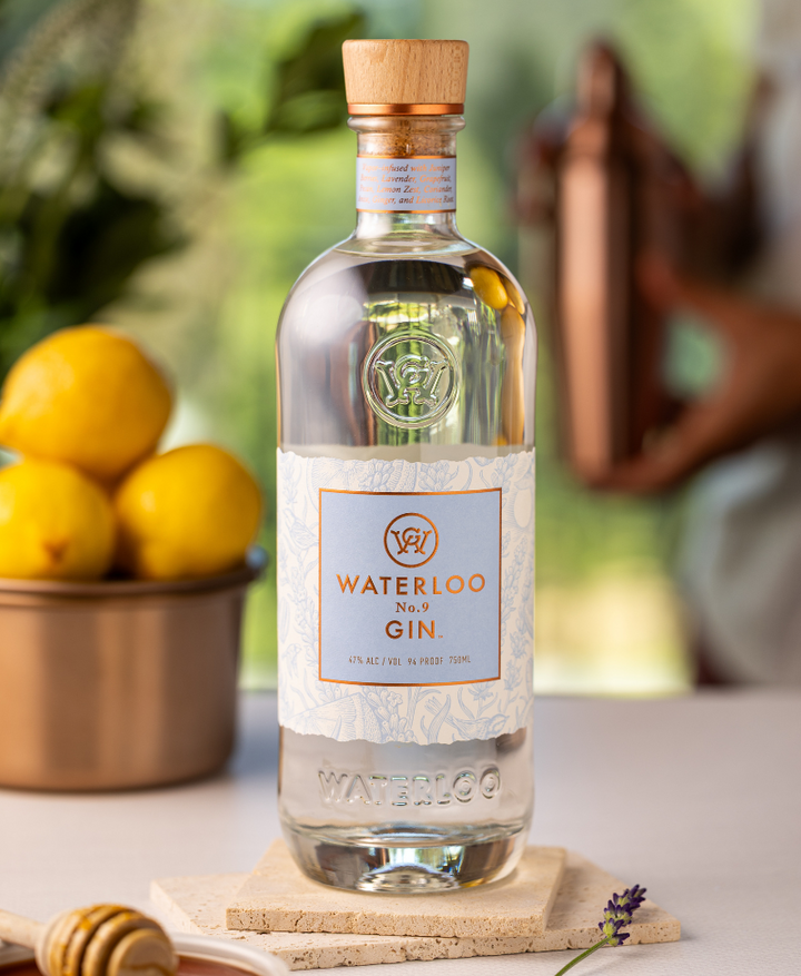 A clear bottle of Waterloo No.9 Gin by Waterloo Gin rests on a coaster. Behind it, lemons, a honey dipper, and someone holding a copper shaker create charm, with hints of greenery in the background.