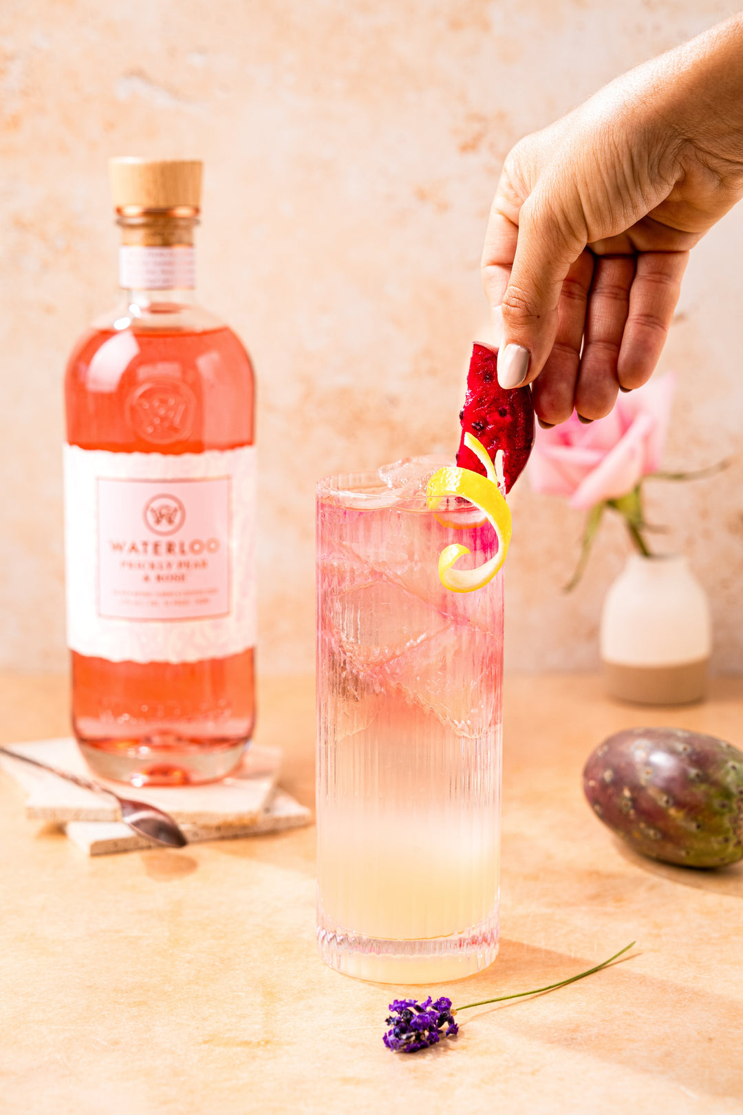 A hand garnishes a tall pink cocktail with dragon fruit and lemon. Behind it stands a pink Waterloo Gin bottle labeled "Waterloo Prickly Pear & Rose Gin," alongside a rose in a vase, spoon, and purple passion fruit on a light surface.