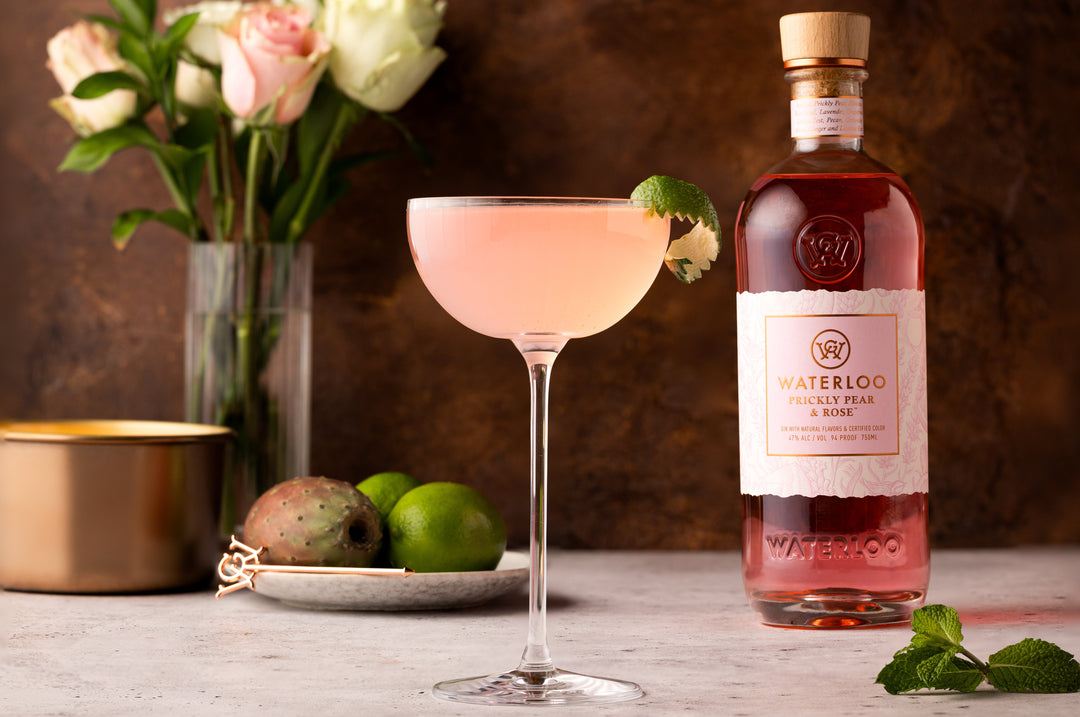 A pink cocktail made with Waterloo Prickly Pear & Rose Gin by Waterloo Gin is garnished with mint in a coupe glass next to the bottle, surrounded by limes, prickly pears, a gold candle, and white roses.