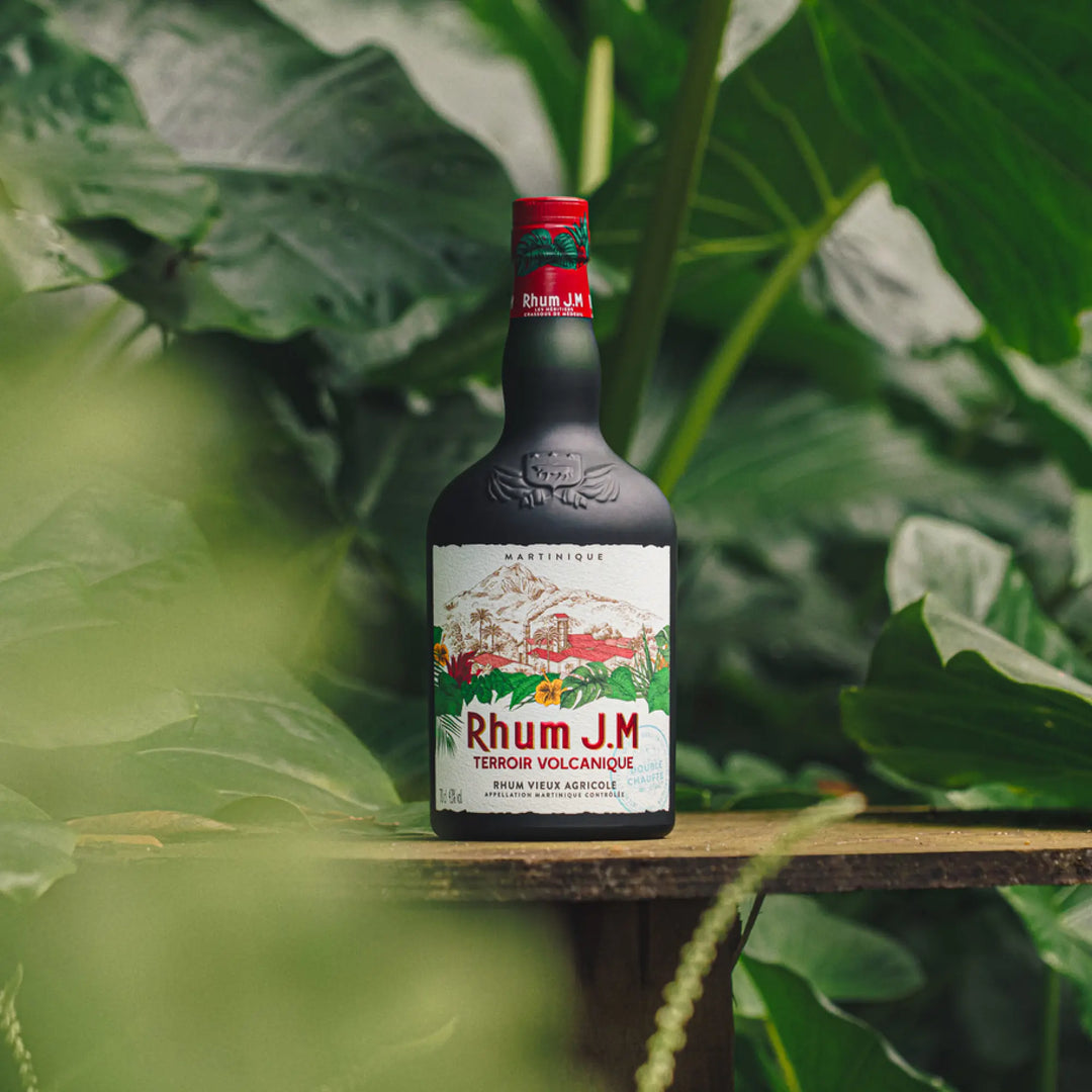 A bottle of Rhum J.M. Terroir Volcanique Rum from Rhum J.M rests on a wooden surface amid lush tropical leaves, its label evoking the double-char process that enhances unique wood flavors and volcanic character.