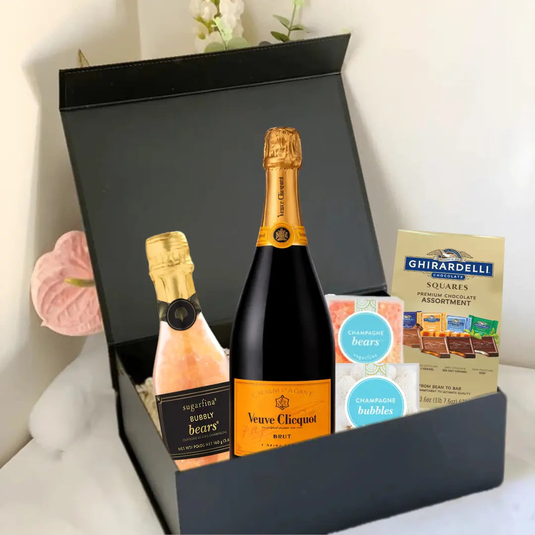 A black Premium Champagne Gift Box by Main Street Liquor sits open on a bed, featuring Veuve Clicquot champagne, Sugarfina bubbly bears, Ghirardelli chocolate squares, and a bath soak—a luxurious champagne gift for any special occasion.