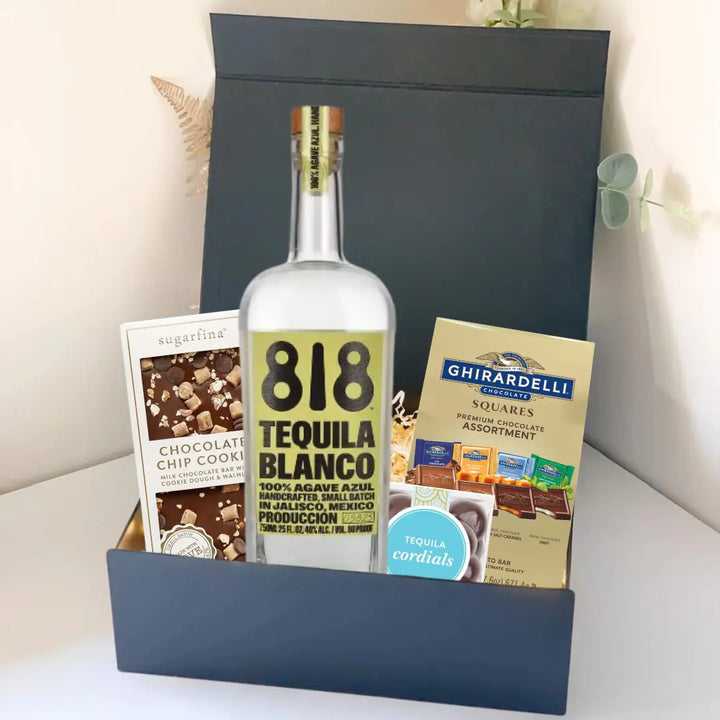 The Premium Tequila Gift Box by Main Street Liquor features 818 Tequila Blanco, Sugarfina chocolate chip cookies, and Ghirardelli squares, beautifully presented on a light surface with a leafy plant in the background.