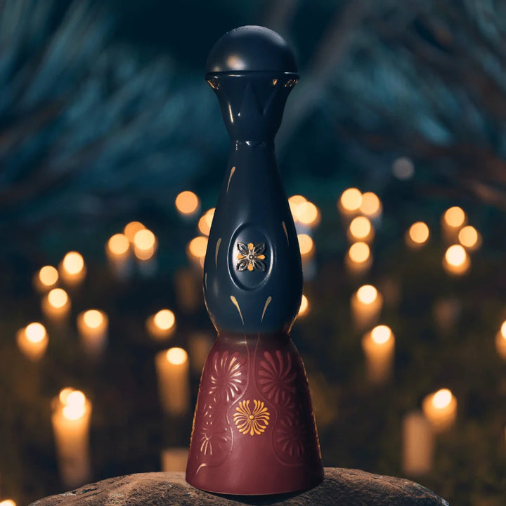 A dark blue and maroon Clase Azul Día de Muertos Trascender 2026 Limited Edition Añejo Tequila 1L decanter with floral engravings, set on a rock against a background of blurred glowing candles.