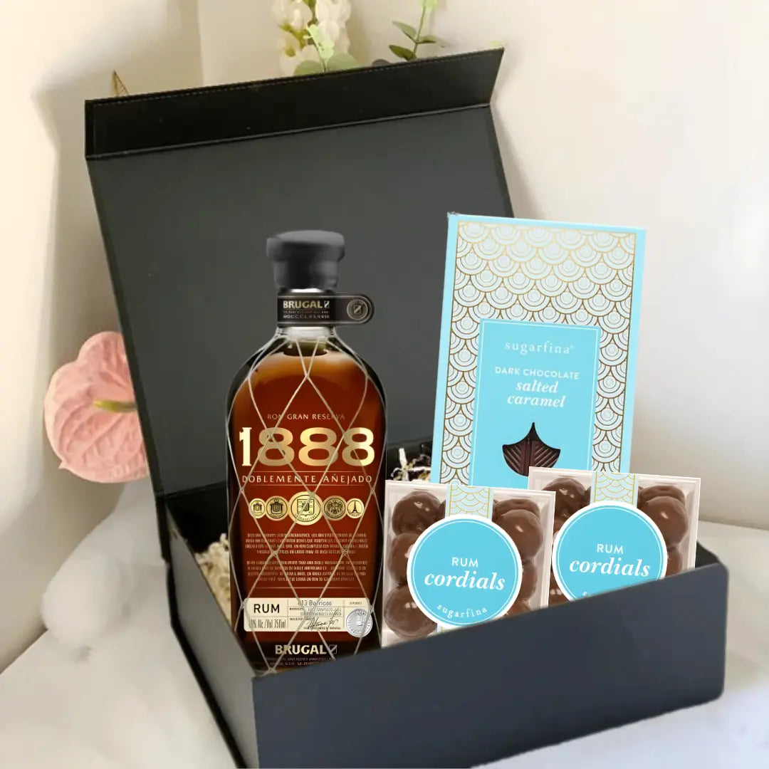 The Main Street Liquor Premium Rum Gift Box includes Brugal 1888, Sugarfina Rum Cordials, dark chocolate sea salted caramels, and two packs of rum cordial chocolates, beautifully arranged on a white surface with a white flower.