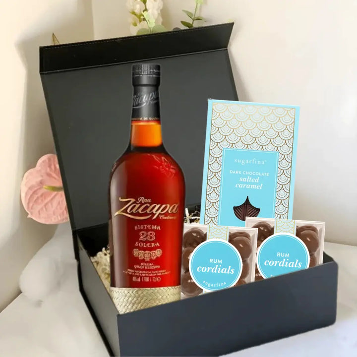 The Premium Rum Gift Box by Main Street Liquor features a bottle of Ron Zacapa, a Sugarfina dark chocolate sea salt caramel bar, and two boxes of Sugarfina Rum Cordials, all presented on white shredded paper with a flower in the background.