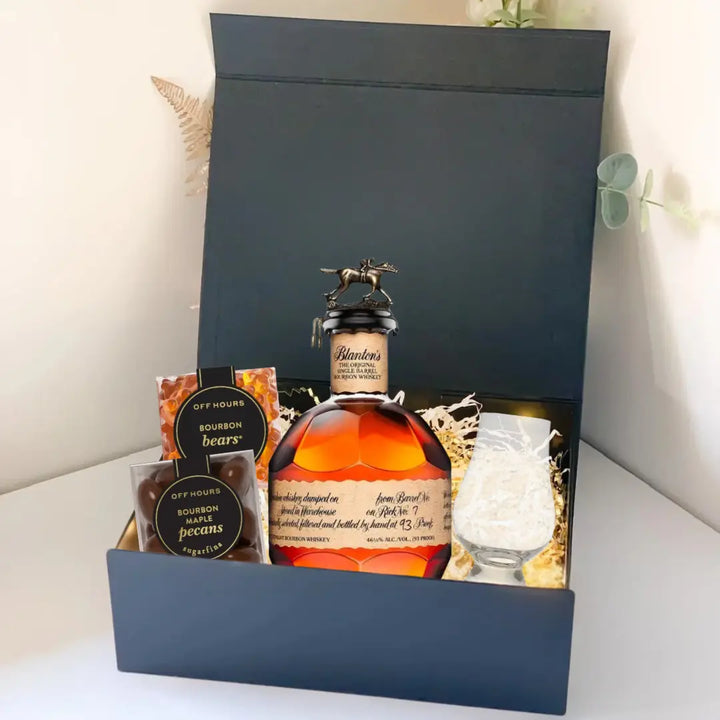 The Premium Bourbon Gift Box by Main Street Liquor includes a bottle of premium bourbon, a whiskey glass, and two packs of bourbon-flavored snacks, all elegantly packed in shredded material inside a sleek black box.