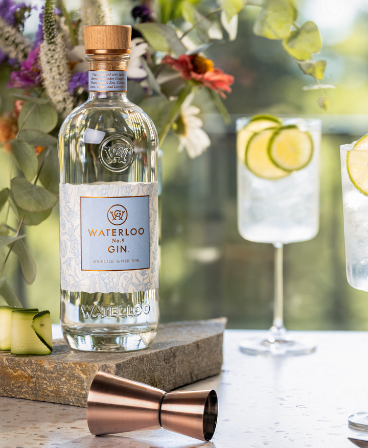 A bottle of Waterloo No.9 Gin by Waterloo Gin rests on a stone slab amid fresh flowers, with two gin and tonics garnished with lime and cucumber behind it, and a copper jigger in front.