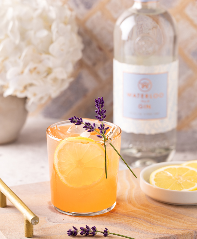 A cocktail garnished with lemon and lavender sits on a wooden board, featuring Waterloo No.9 Gin by Waterloo Gin, with extra lemon slices and white flowers in the background—a fresh taste of Texas Hill Country.