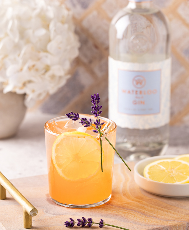 A cocktail garnished with lemon and lavender sits on a wooden board, featuring Waterloo No.9 Gin by Waterloo Gin, with extra lemon slices and white flowers in the background—a fresh taste of Texas Hill Country.