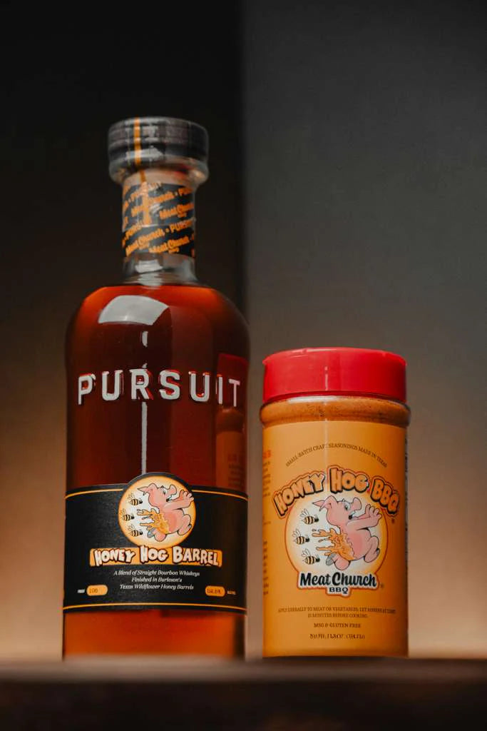 A bottle of Pursuit Spirits Pursuit x Meat Church Honey Hog Barrel Bourbon stands next to Meat Church Honey Hog BBQ seasoning, both displayed on a dark background with warm lighting.