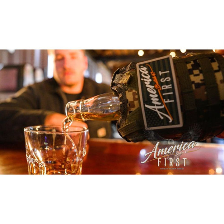 A close-up of the America First Straight Bourbon Whiskey 3 Pack wrapped in a camouflage sleeve pours bourbon into a glass, while a blurred man sits at a wooden bar—a unique addition for any America First collector.