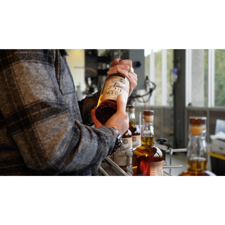 Wearing a plaid jacket, a person holds a bottle from the America First Straight Bourbon Whiskey 6 Pack at a counter with other America First bottles nearby and large windows in the background.