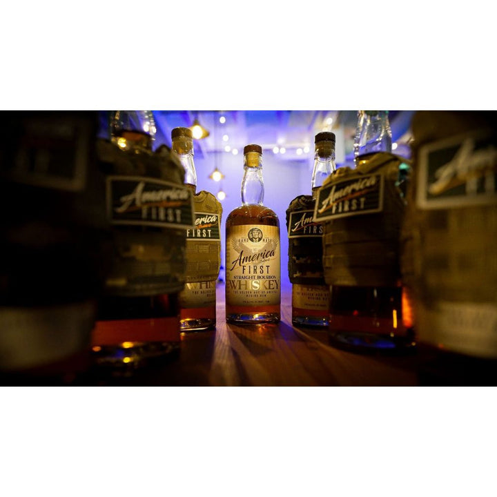 A 2-pack of America First Straight Bourbon Whiskey by America First sits on a table, surrounded by a bourbon collector’s set, with string lights and warm ambient lighting in the background.