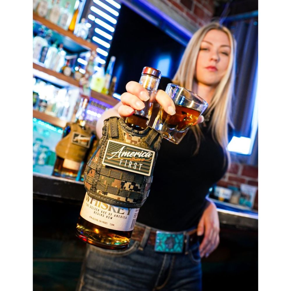 A woman at a bar holds a glass of whiskey and an America First Straight Bourbon Whiskey 6 Pack bottle wrapped in camouflage, with bottles and shelves visible behind her.