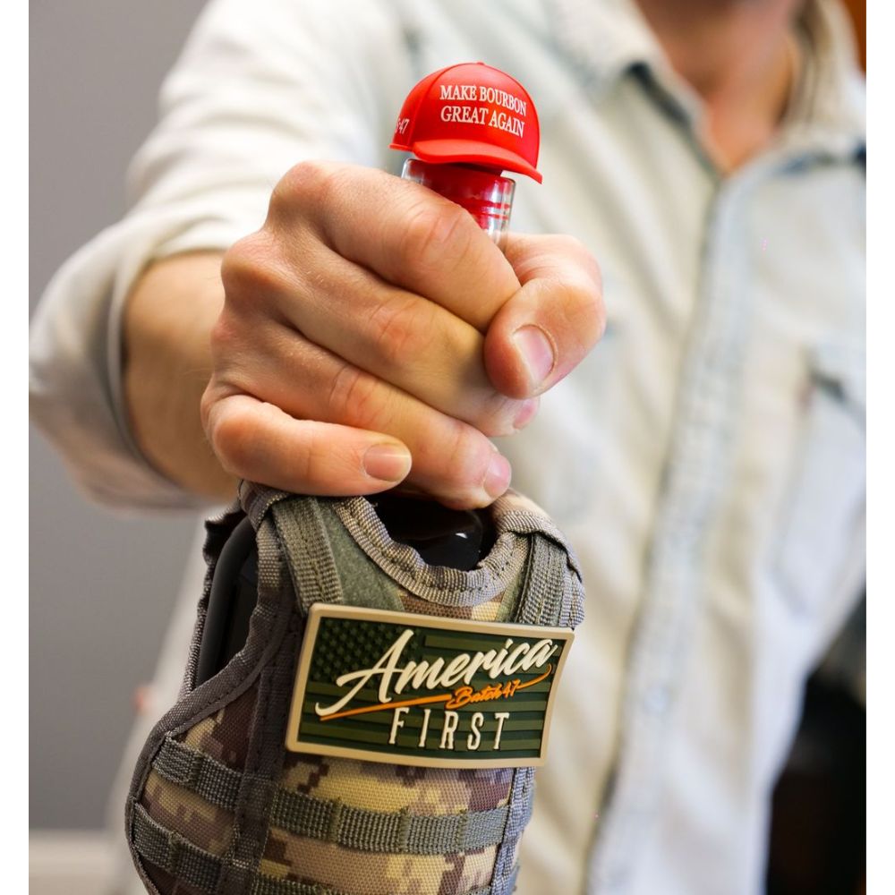 A person holds a bottle of America First Straight Bourbon Whiskey with a red cap labeled "MAKE BOURBON GREAT AGAIN." The bottle, from the America First brand, is in a camouflage sleeve with an America First patch. Only their hand and torso are visible.
