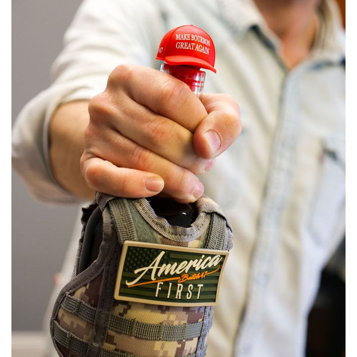 A person holds a bottle of America First Straight Bourbon Whiskey with a red cap labeled "MAKE BOURBON GREAT AGAIN." The bottle, from the America First brand, is in a camouflage sleeve with an America First patch. Only their hand and torso are visible.