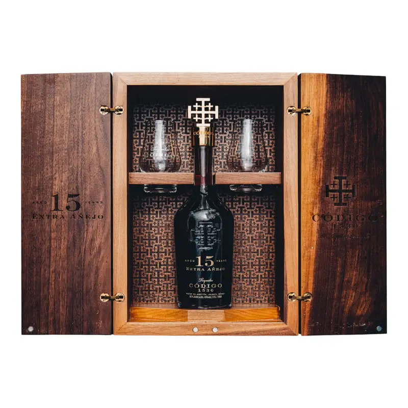 A wooden box with engraved designs showcases the Código 1530 Tequila Extra Añejo 15 Year Gift Set, featuring a centered bottle below two glass tumblers on a shelf, all set against a patterned interior background.