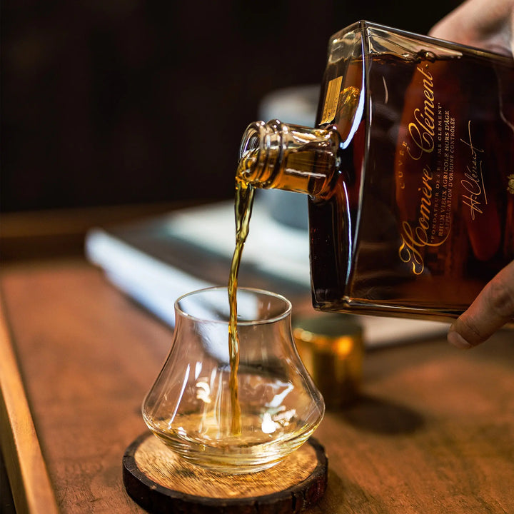 A hand pours amber Rhum Clément Cuvée Homère from a square bottle into a curved glass on a wooden coaster, with a blurred book in the background.