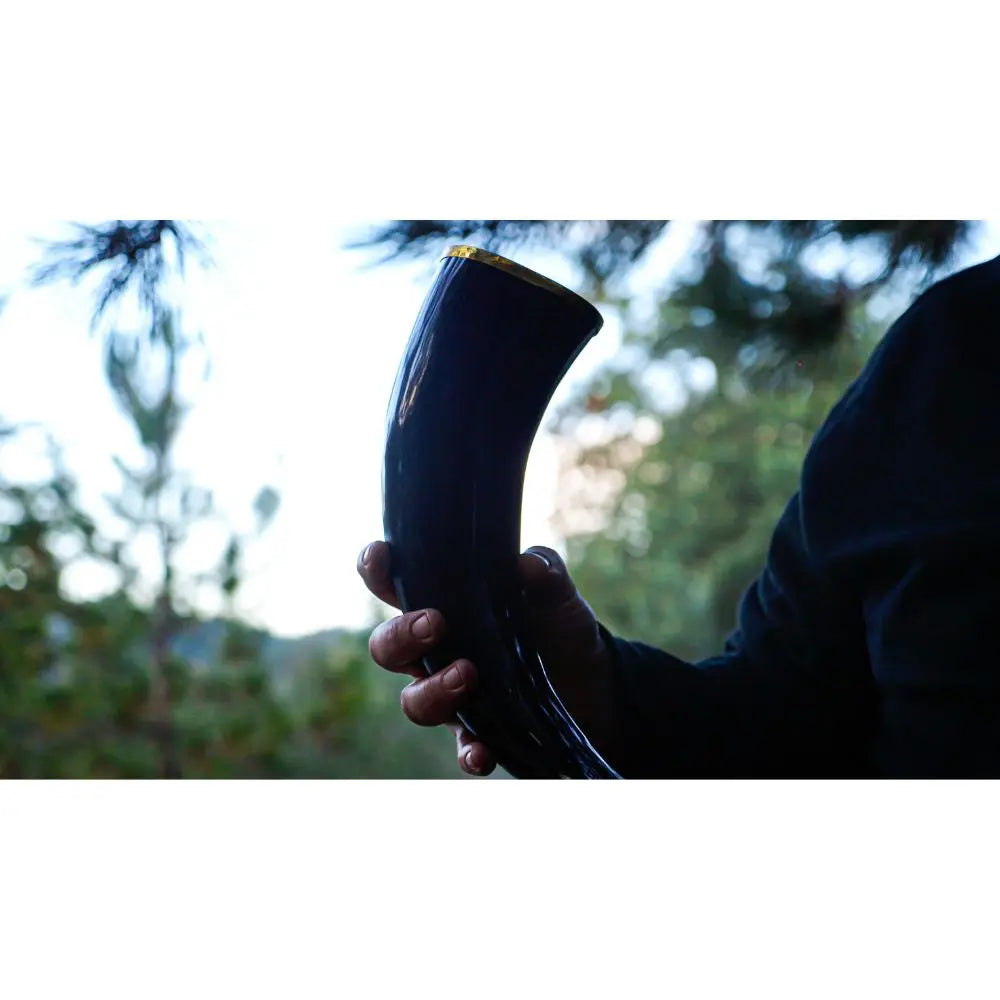 A person in a dark sleeve holds a polished drinking horn outdoors, savoring the moment as sunlight hits the greenery—perhaps toasting with Eldenwood Straight Bourbon Whiskey from Eldenwood among the blurred trees.