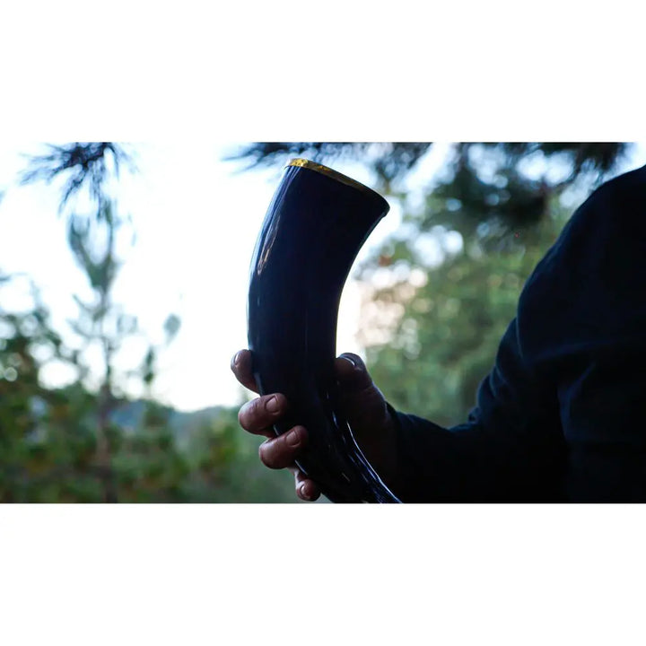 A person in a dark sleeve holds a polished drinking horn outdoors, savoring the moment as sunlight hits the greenery—perhaps toasting with Eldenwood Straight Bourbon Whiskey from Eldenwood among the blurred trees.