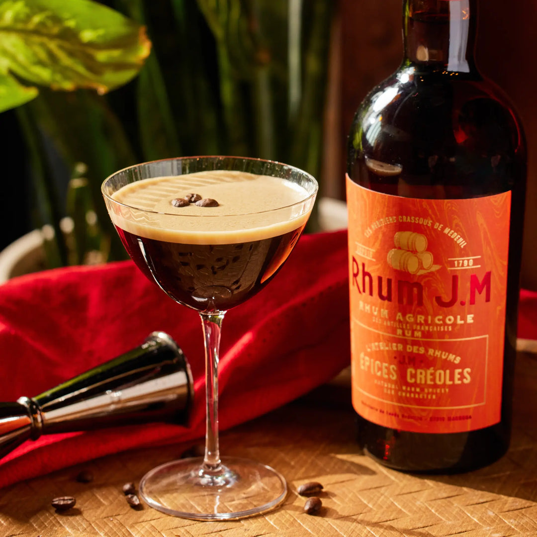A cocktail glass of frothy coffee, topped with coffee beans, sits beside a bottle of Rhum J.M Atelier Épices Créoles on a wooden surface, with a red cloth, metal bar tool, and green plants in the background.