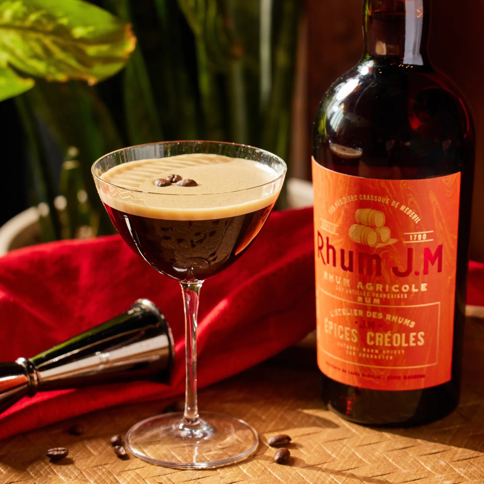A cocktail glass of frothy coffee, topped with coffee beans, sits beside a bottle of Rhum J.M Atelier Épices Créoles on a wooden surface, with a red cloth, metal bar tool, and green plants in the background.