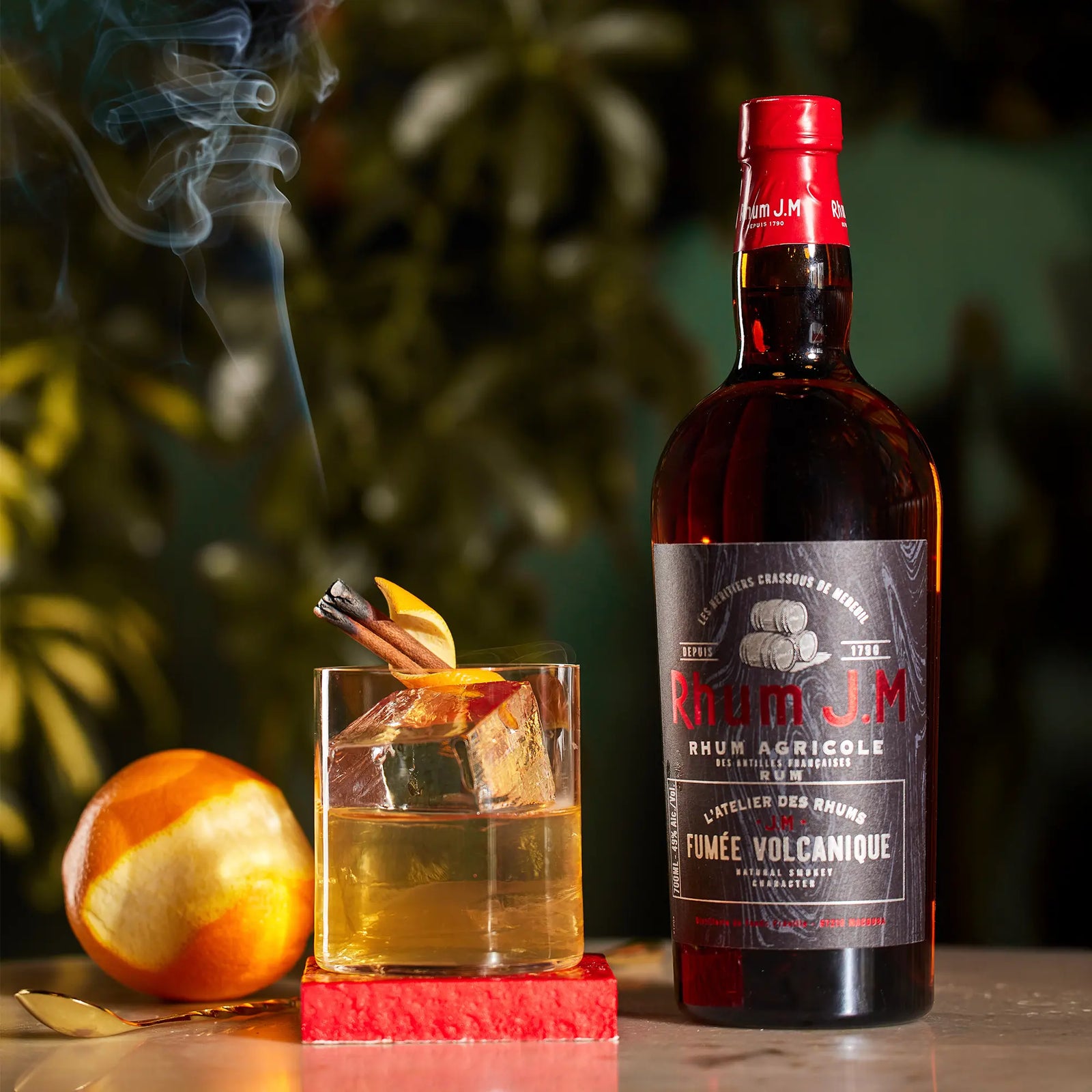 A bottle of Rhum J.M Atelier Fumée Volcanique stands by a smoky cocktail with cinnamon stick and orange peel garnish, as an orange sits nearby on the table.