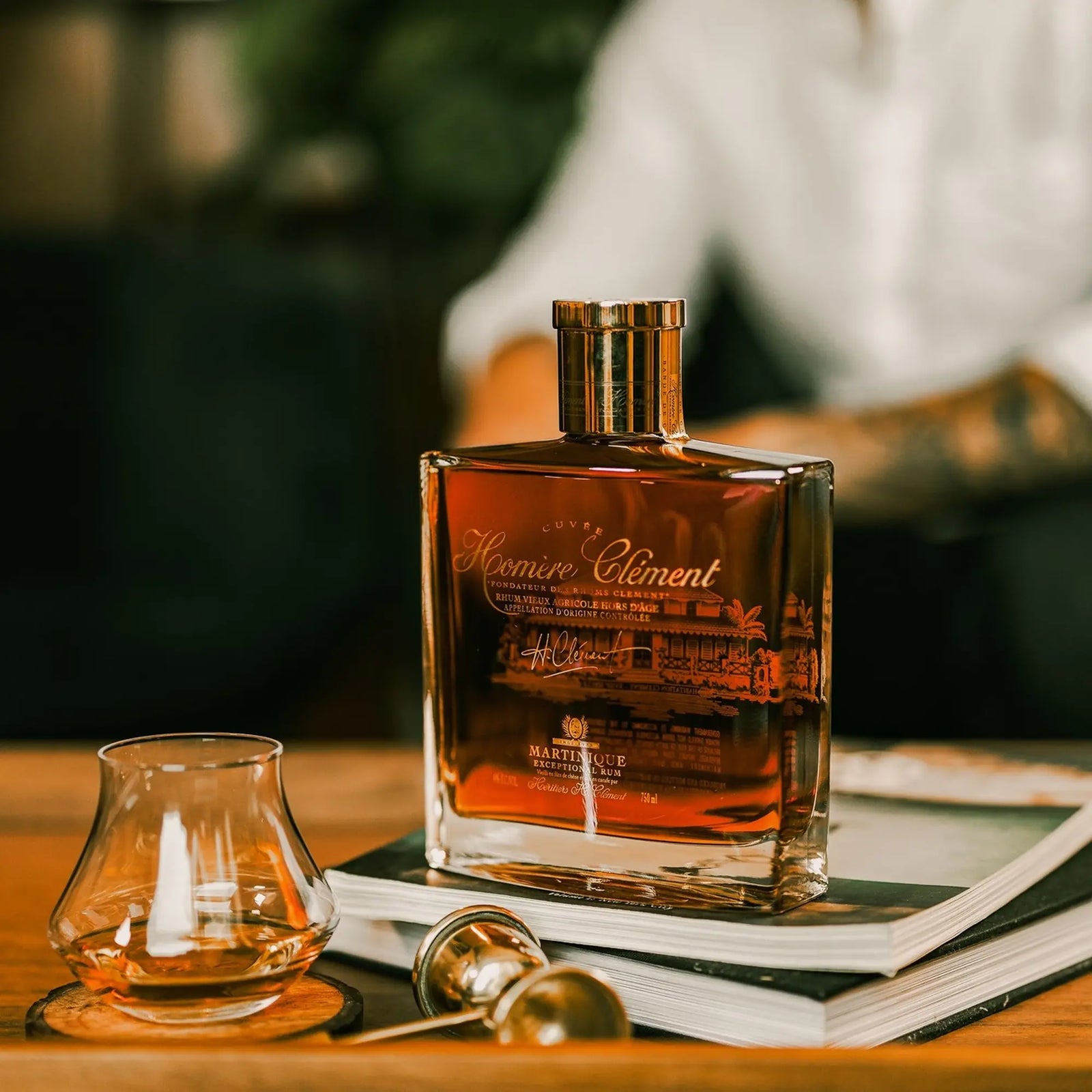 A square bottle of Rhum Clément Cuvée Homère sits on books beside a glass of rhum agricole and a metal jigger on a wooden table, with a blurred figure in a white shirt in the background.