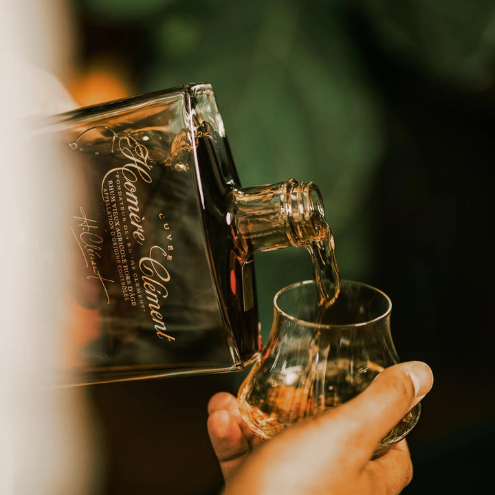 A close-up of a hand pouring amber Rhum Clément Cuvée Homère from a square glass bottle into a rounded whiskey glass, with a blurred green background.