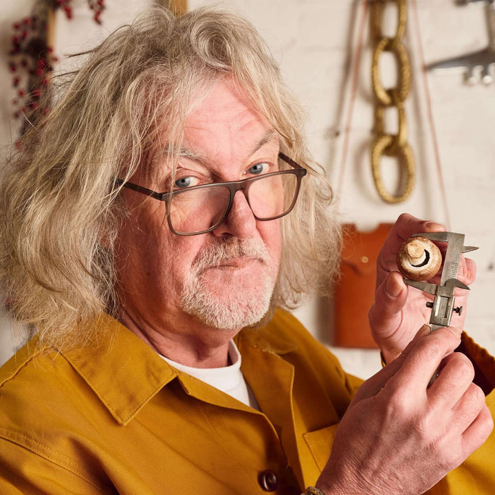 An older man with gray, wavy hair and glasses, in a mustard shirt, measures a small object in his workshop. Tools and chains hang behind him, capturing the inventive spirit of James Gin's California Dreamgin' with Souvenir Gift Key Chain.