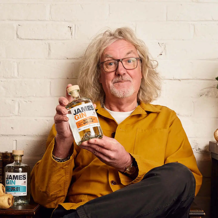 A gray-haired man with glasses in a yellow jacket sits against a white brick wall, smiling and holding a bottle of James Gin California Dreamgin' W/ Souvenir Gift Key Chain. Another James Gin bottle is on the table next to him.