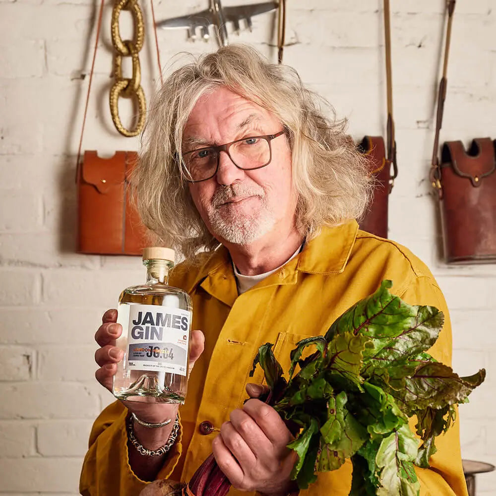 A man with long gray hair and glasses, wearing a yellow jacket, stands by a white brick wall with leather bags behind him, holding James Gin London Drizzle 700mL 80 Proof W/ Souvenir Gift Key Chain in one hand and leafy greens in the other.