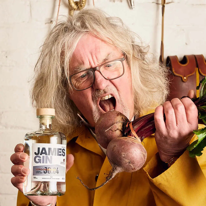 A man with long, messy gray hair and glasses in a yellow coat holds a bottle of James Gin London Drizzle 700mL 80 Proof W/ Souvenir Key Chain in one hand and beetroots in the other, appearing to shout or bite the beetroots.