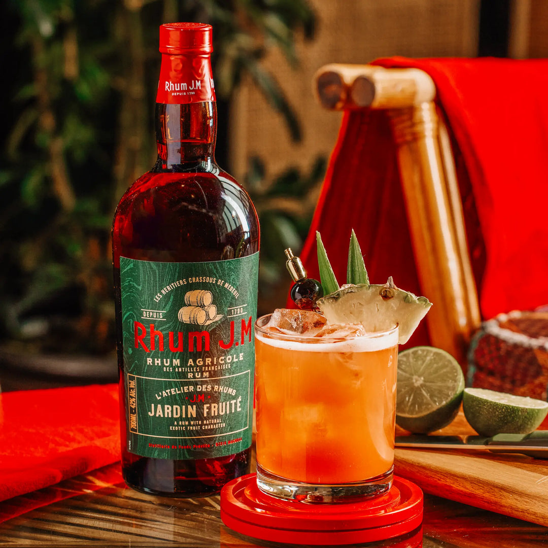 A bottle of Rhum J.M Atelier Jardin Fruité by Rhum J.M stands next to a colorful cocktail topped with pineapple, lime, and leaves on a red coaster, with lime halves and a wooden muddler nearby in this tropical scene.