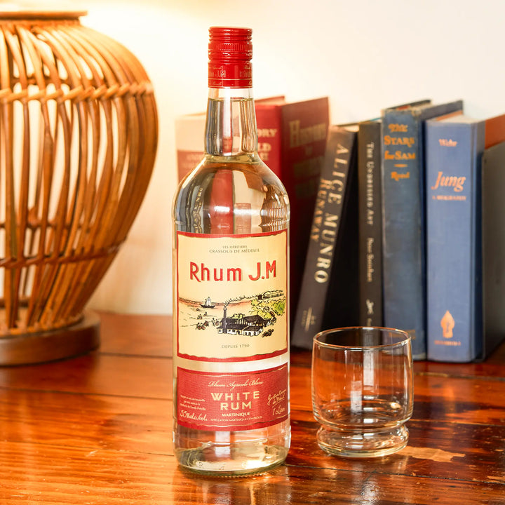 A bottle of Rhum J.M. Agricole Blanc 110 Proof White Rum stands on a wooden table beside an empty glass, with books and a wicker lamp in the background.