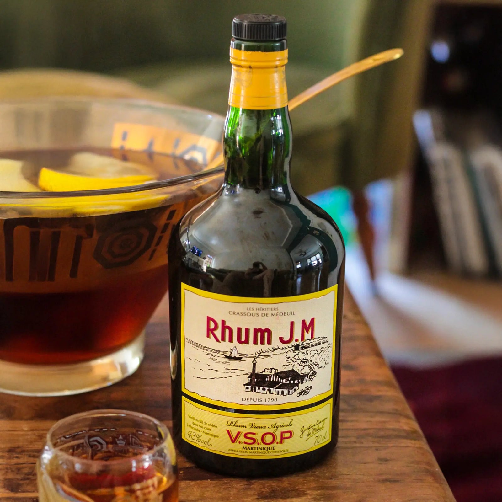 A bottle of Rhum J.M. V.S.O.P. Martinique Aged Rum sits on a wooden table beside a glass punch bowl with lemon slices and a small glass of dark rum. Its yellow label features red and black text—crafted by Rhum J.M in Bourbon barrels.