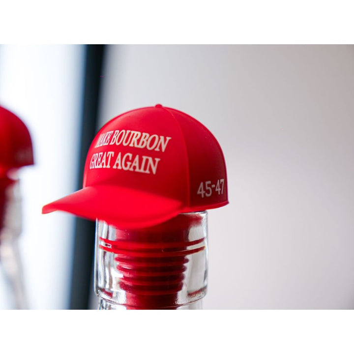 A red "MAKE BOURBON GREAT AGAIN" 45-47 baseball cap tops an America First Straight Bourbon Whiskey bottle, serving as a festive bottle topper and showcasing American craftsmanship.