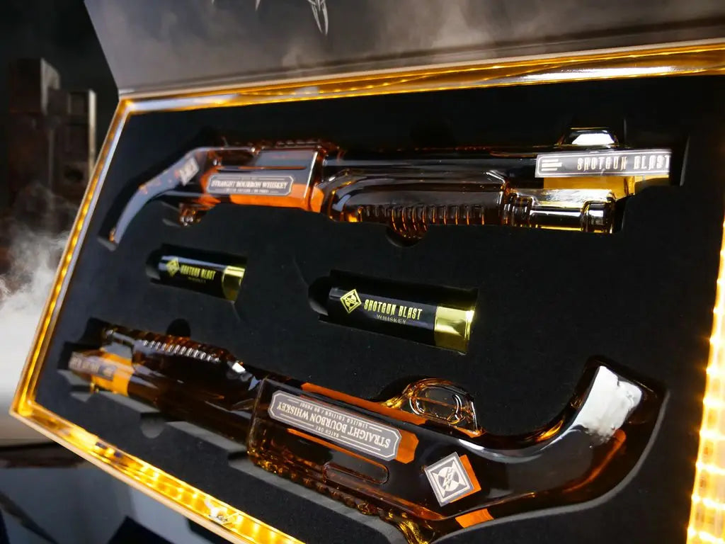 Two glass bottles shaped like shotguns, labeled Shotgun Blast Whiskey First Edition Straight Bourbon By Machine Head, are displayed in a collector’s box with foam lining, alongside two gold-colored cylindrical containers. Brand: Shotgun Blast.