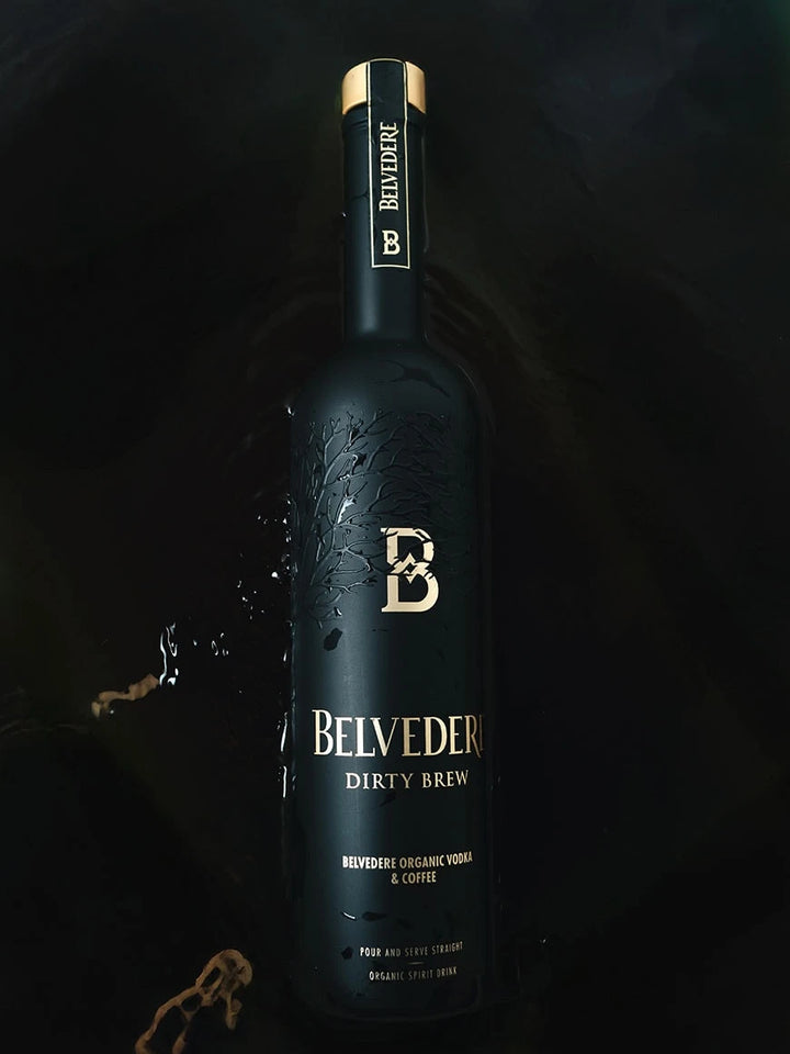 A matte black bottle of Belvedere Dirty Brew Organic Coffee Flavored Vodka by Belvedere Vodka, accented with gold lettering, rests on a dark surface amid coffee splashes. The label highlights “Organic Vodka &amp; Coffee.”.