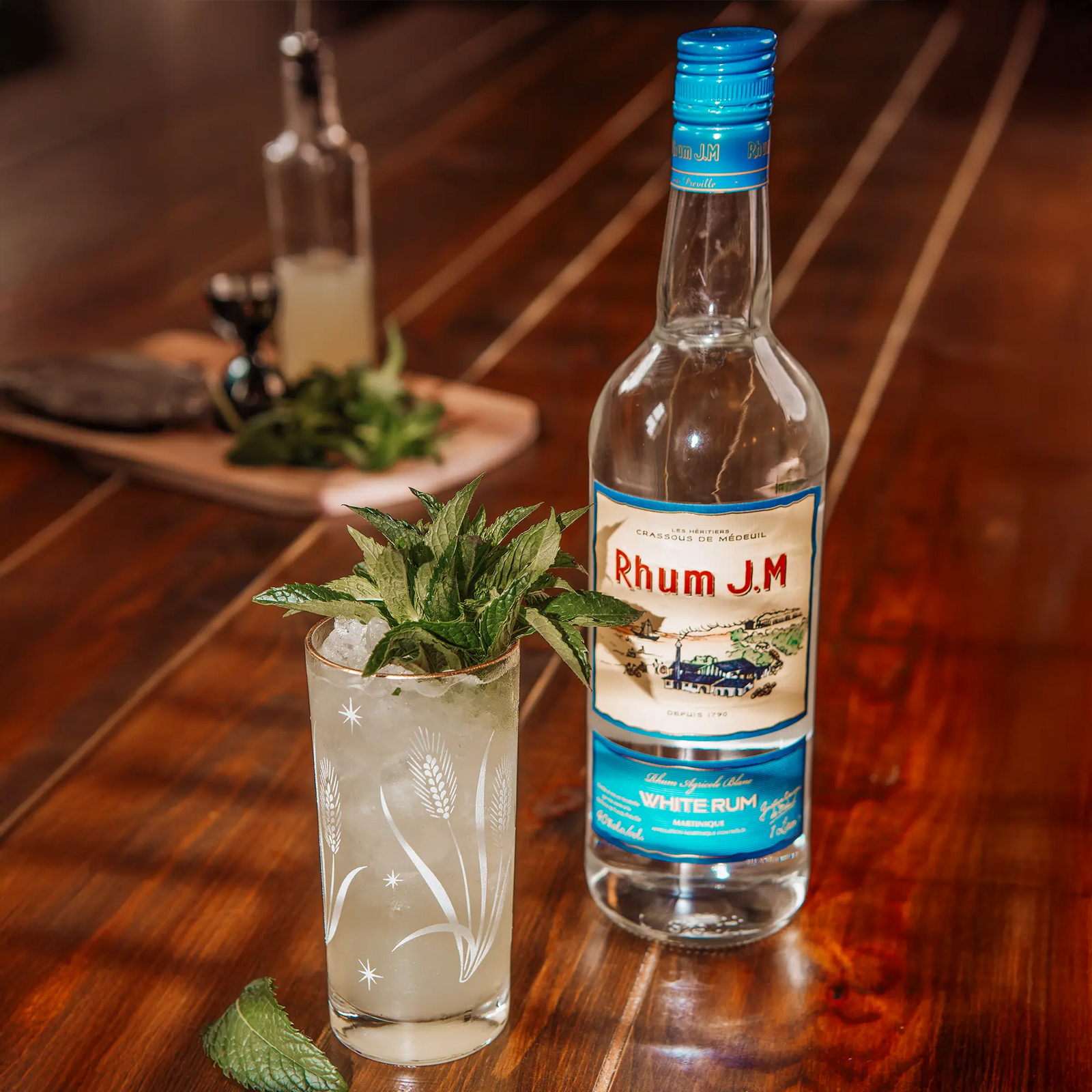 A bottle of Rhum J.M. Agricole Blanc 40 White Rum sits beside a mint-garnished cocktail on a wooden table, offering a refined experience with blurred ingredients and hints of grilled pineapple in the background.