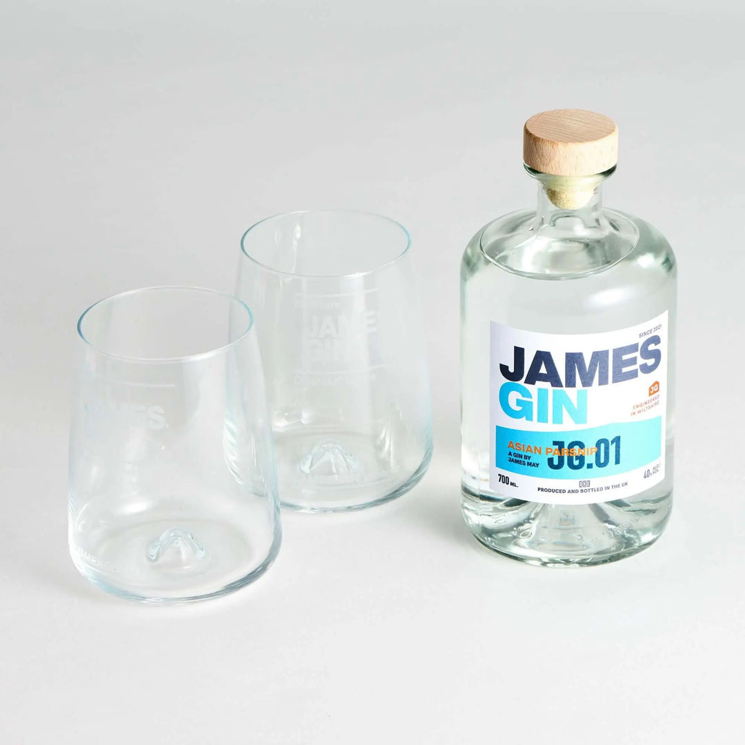 A James Gin The Sharing Cocktail Kit W/ Souvenir Gift Key Chain by James Gin US sits beside two empty clear glasses on a white surface. The bottle has a wooden cap and features a white and blue label with text and numbers.
