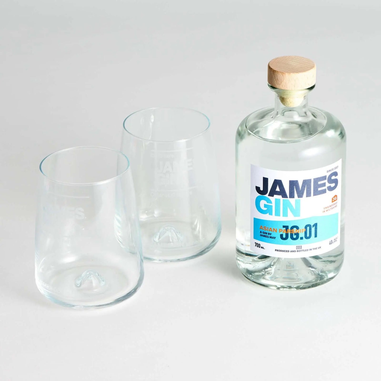 A James Gin The Sharing Cocktail Kit W/ Souvenir Gift Key Chain by James Gin US sits beside two empty clear glasses on a white surface. The bottle has a wooden cap and features a white and blue label with text and numbers.